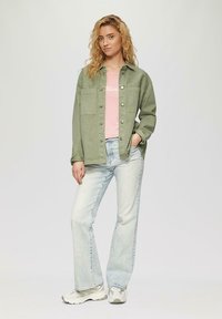 Veste en denim vert olive sur un t-shirt rose clair, associée à un jean bleu clair et à des baskets beiges. Coupe standard et style décontracté.