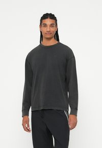 Tamno siva duga rukava dukserica s okruglim izrezom i opuštenim krojem. Mekan materijal s vidljivim šavovima. Nosena s crnim hlačama.