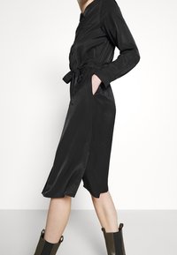 Robe en satin noir avec une taille ceinturée, poches latérales, manches longues et une longueur au genou, dotée d'une fermeture à boutons et d'un design épuré.