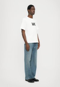 Hvit bomull T-skjorte med svart MK-logo, sammen med lyseblå jeans. Modellen står i profil foran en ensfarget bakgrunn. Svart sko.
