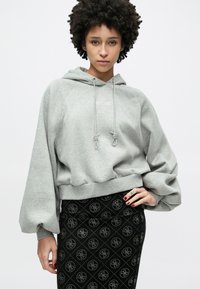 Γκρι cropped φούτερ με κουκούλα και κορδόνι, υφασματικό σώμα και κεντημένο «EST. 1981.» Συνδυάζεται με μαύρη φούστα με σχέδιο και λογότυπα.