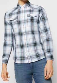 Camicia a maniche lunghe a quadri in tonalità blu e nero, con colletto abbottonato, tasche sul petto e dettagli di cuciture bianche. Materiale in misto cotone.