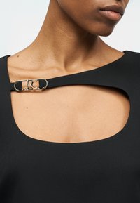 Κοντινό πλάνο ενός ατόμου που φοράει μαύρο τοπ με φαρδύ ντεκολτέ και χρυσή λεπτομέρεια κλεισίματος στον αριστερό ώμο.