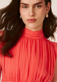 Mujer con cabello oscuro lleva blusa sin mangas de cuello alto color coral con pliegues y pendientes blancos en forma de lágrima, fondo neutro.