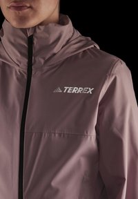 Voděodolná bunda s kapucí v prachově růžové barvě, s uzavíráním na zip a reflexním logem "TERREX" na hrudi s černými akcenty.