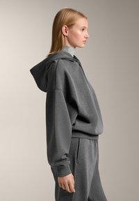 Siva majica s kapuljačom i spuštenim ramenima s džepom na trbuhu, izrađena od mekanog materijala. Ima rebraste manžete i opušteni kroj.