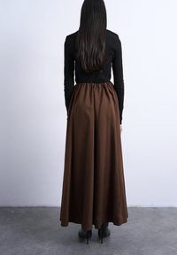 Γυναίκα με μακριά σκούρα μαλλιά, που φοράει μαύρο μακρυμάνικο τοπ, καφέ ψηλόμεση παντελόνα με φαρδιά πόδια και μαύρες μυτερές γόβες, στέκεται μπροστά σε έναν λευκό τοίχο.