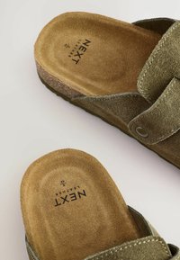 Olive green suede sandals with a circular toe shape, adjustable strap, and textured insole featuring "NEXT Leather" branding. Cork-like sole.
