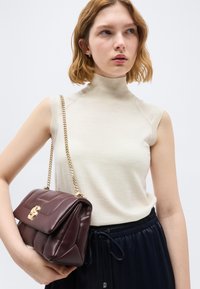 Νεαρή γυναίκα φοράει μπεζ ζιβάγκο χωρίς μανίκια και μαύρο παντελόνι, κρατώντας καφέ σκούρη δερμάτινη τσάντα χειρός με χρυσή αλυσίδα.