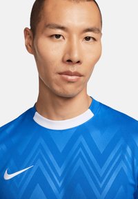 Blauwe sporttrui met een contrasterende witte kraag, voorzien van een zigzagpatroon over de stof. Inclusief een klein wit Nike-logo op de borst.