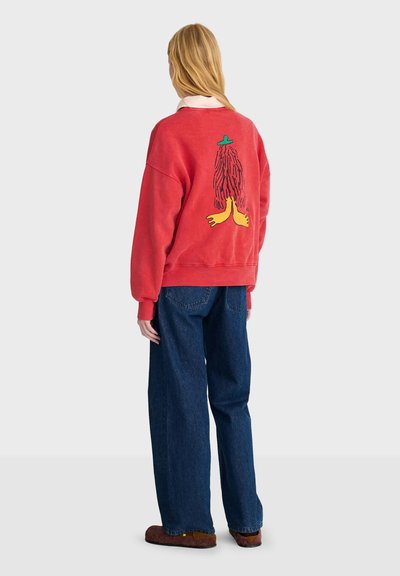 Personne aux longs cheveux blonds portant un sweat rouge avec un motif abstrait coloré dans le dos, un jean bleu et des chaussures marron, debout de dos.
