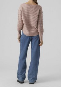 Mujer vestida con un suéter de punto texturizado rosa y vaqueros de pierna ancha azules, de pie mirando hacia atrás sobre un fondo gris liso.