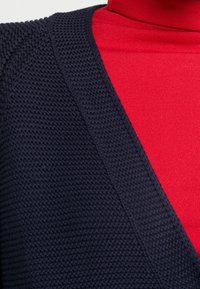 Maglione lavorato a maglia blu scuro con scollatura a V, indossato sopra un dolcevita rosso brillante. Il tessuto testurizzato crea un contrasto visivo.