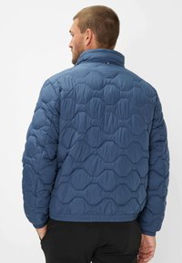 Blå quiltet jakke med høj krave og elastiske manchetter. Har et tekstureret sekskantet mønster, designet til varme og komfort.