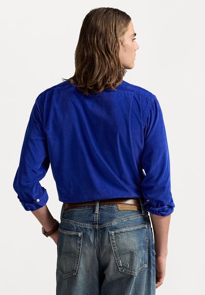 Mann med skulderlangt hår iført en lys blå langermet skjorte og falmede blå jeans, som står vendt bort med hendene nær lommene.