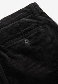 I pantaloni in velluto a coste neri presentano un tessuto testurizzato con righe verticali, una tasca posteriore con un singolo bottone nero e una vita piatta.