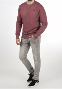 Red knit sweater with a crew neck and black logo detail, paired with light gray denim jeans and black high-top sneakers.