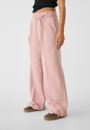 Woman wearing high-waist wide-leg pink trousers with a tied belt, paired with brown sneakers and a cropped black ribbed top.