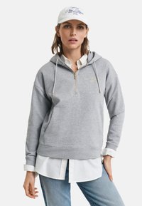 Naine kannab valget mütsi, halli poolepika tõmblukuhoodie't valge särgi peal ja siniseid teksaseid, seistes ühe käega taskus valge tausta vastas.