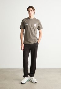 Light olive green cotton t-shirt with printed text and a pink flower design, paired with black pants and white sneakers with green accents.