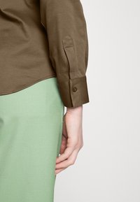 Camicia marrone a maniche lunghe con dettagli sui polsini, abbinata a pantaloni verdi con texture. Focus sulla texture del tessuto, sull'accento dei bottoni e sul contrasto di colori.