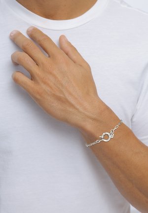 Bracelet chaîne argentée avec fermoir rond, accents circulaires et petit élément décoratif. Porté sur un poignet bronzé, avec un T-shirt blanc uni.