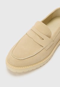 Beige Wildlederschuh zum Hineinschlüpfen mit einer erhöhten Naht, einem dekorativen Riemen über der Vorderseite und einer strukturierten Gummisohle. Glatte Innenauskleidung.