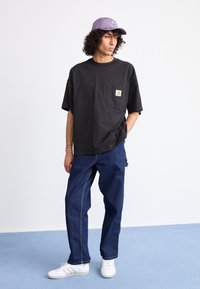 Young person wearing a black oversized T-shirt, blue jeans, white sneakers, and a purple Adidas cap, standing on a blue floor against a white wall.