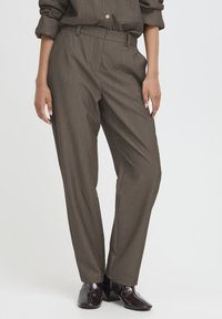 Braune, maßgeschneiderte Hose mit geradem Bein, feiner Textur und Gürtelschlaufen, kombiniert mit dunklen, glänzenden Stiefeletten.