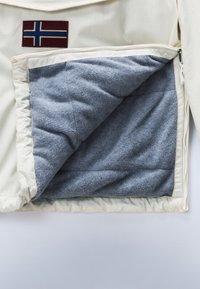 Chaqueta blanca con forro polar gris parcialmente visible, con un parche de cruz nórdica en azul marino y granate en la parte superior izquierda del pecho.