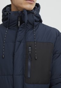 Chaqueta acolchada de color azul marino con cuello alto, que presenta un bolsillo con cremallera negra en contraste y detalles de cordón. El material parece suave.