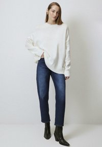 White oversized sweater with ribbed cuffs, paired with dark blue jeans and black heeled ankle boots. Model stands against a neutral background.