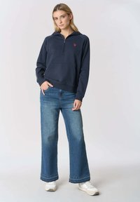 Felpa con zip blu navy con tasca frontale e logo rosso, abbinata a jeans blu a gamba larga e sneakers bianche. Tessuto dalla texture liscia.