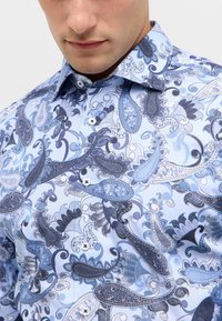 Camicia blu con motivo paisley, caratterizzata da bottoni bianchi e un colletto strutturato, realizzata in un tessuto morbido con dettagli intricati in diverse tonalità di blu.