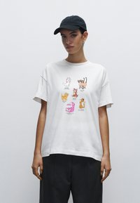 Hvid bomulds t-shirt med cartoon-katte i forskellige positioner og farver, med en afslappet pasform og korte ærmer. Sæt sammen med mørke bukser.