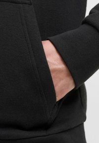Schwarzer Hoodie mit glatter Textur, ausgestattet mit einer Kängurutasche und gerippten Bündchen. Nahaufnahme einer Hand, die in der Tasche ruht.