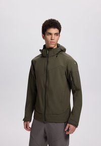 Jaunas vyras dėvi alyvuogių žalios spalvos gobtuvu apvilktą striukę su užtrauktuku, stovi priešais šviesų, vientisą foną, žiūri šiek tiek į dešinę.