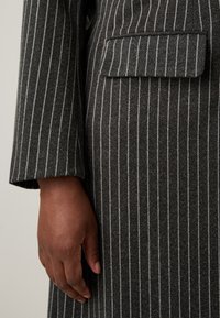 Sivor prugast plašč iz teksturirane tkanine, ki ima en sam žep na prsih in prilagojene rokave. Roka, ki počiva na robu plašča.