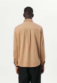 Chemise marron à manches longues avec un col, des poignets à bouton et un dos plissé, fabriquée en matériau doux ressemblant au coton.