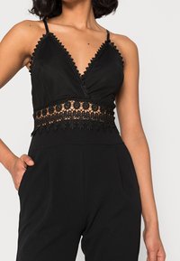 Kvinna i en svart ärmlös jumpsuit med spetsdetaljer och virkad midja, poserar med en hand i fickan mot en neutral bakgrund.