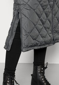 Gonna grigia trapuntata con zip laterale indossata sopra leggings neri e stivali alla caviglia in pelle nera con lacci su un pavimento grigio.
