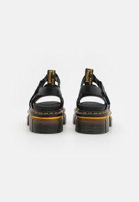 Svarta plattformssandaler med en texturerad ovandel, öppen design, gul och svart branding på hälen, samt en lagerad sula med upprepande mönster.