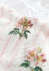 Primo piano di tessuto bianco con motivi testurizzati, stampe floreali rosa e un piccolo charm rotondo in metallo dorato attaccato vicino al bordo superiore.