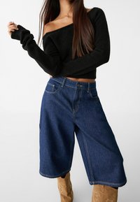 Zwarte off-the-shoulder gebreide trui met geribbelde manchetten, gecombineerd met donkerblauwe denim wijde culotten met zichtbare stiksels en zakken.