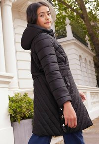 Abrigo acolchado negro con capucha, que presenta un diseño acolchado, detalles de botones y una cintura ceñida, llevado sobre unos vaqueros azules.