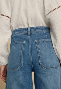 Vaqueros de denim azul de tiro alto con dos bolsillos traseros, una silueta ajustada y detalles de costura sutiles. Combinados con una blusa blanca texturada.