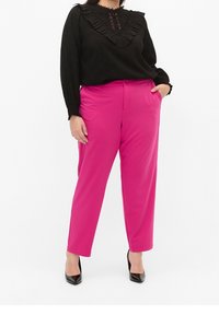Haut noir à manches longues avec encolure à volants et détails en dentelle, associé à un pantalon tailleur rose vif. Escarpins noirs à bout pointu.