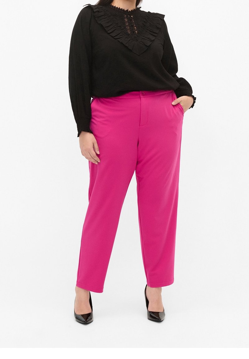 Haut noir à manches longues avec encolure à volants et détails en dentelle, associé à un pantalon tailleur rose vif. Escarpins noirs à bout pointu.