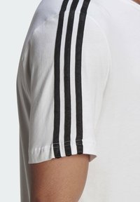 Witte T-shirt met korte mouwen, voorzien van drie zwarte strepen langs de schouder, met een zachte textuur en een lichte glans op de stof.