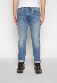 Világoskék farmernadrág slim fit szabással, felgöngyölt szárakkal és öt zsebes kialakítással. Fekete és narancssárga sportcipőkkel párosítva.